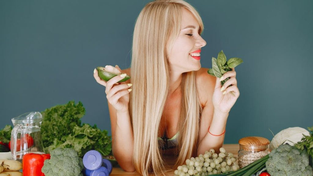 A smiling woman holding avocado and basil, surrounded by fresh vegetables and fruits for healthy eating.